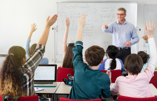 Teacher standing in from of class with students raising hands to be called on