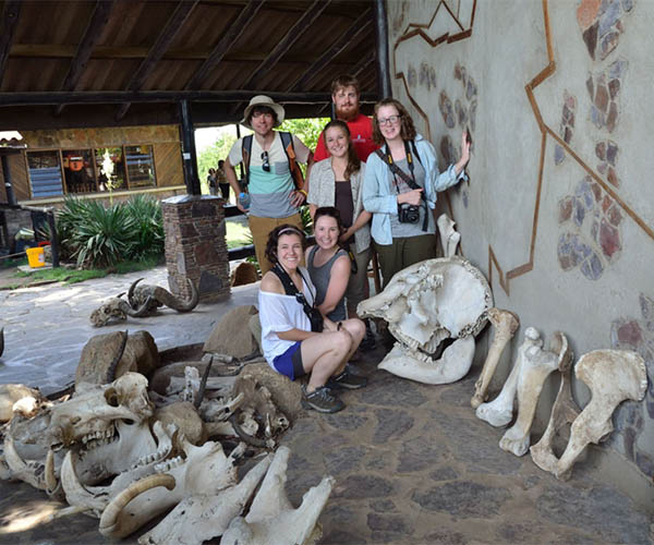 Six students, some with cameras, smiling and posing next to large animal fossils