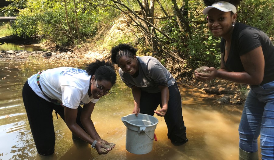 Three female students putting sand into a bucket in a creek