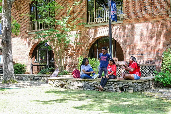 Three students collaborating at a laptop in a bright campus study area, focused and engaged, surrounded by books and modern furniture