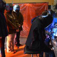 Three individuals watching a metal worker, all in protective gear 
