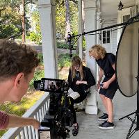 - 

Behind-the-scenes film shoot on a porch, with a camera operator filming a person standing near a reflector.