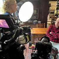 Close-up of a film camera with a bright circular light, with a person in a red shirt visible in the background near a wooden dresser.
