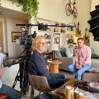 Interview setup inside a home, with two people seated across from each other and a camera on a tripod in the foreground.