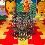 Large spiky metal sculpture on colorful checkered floor between painted totems
