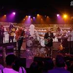 Wide shot of a full band performing on a large stage with purple lighting and an audience in the foreground.