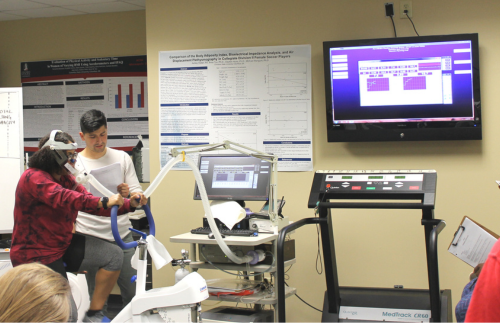 A lady riding a stationary bike hooked up to a breathing machine with a man monitoring her