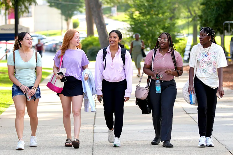 Five college students walking together and smiling on a sunny campus sidewalk