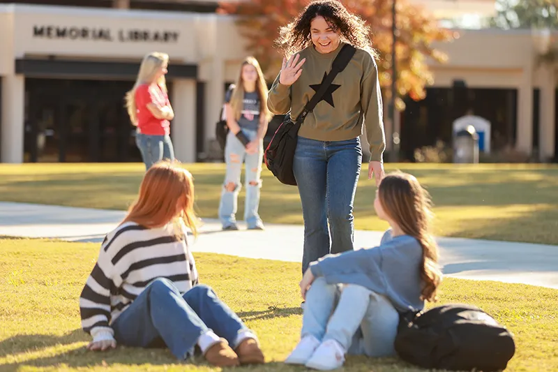 A student waving to two friends sitting on the grass outside the library
