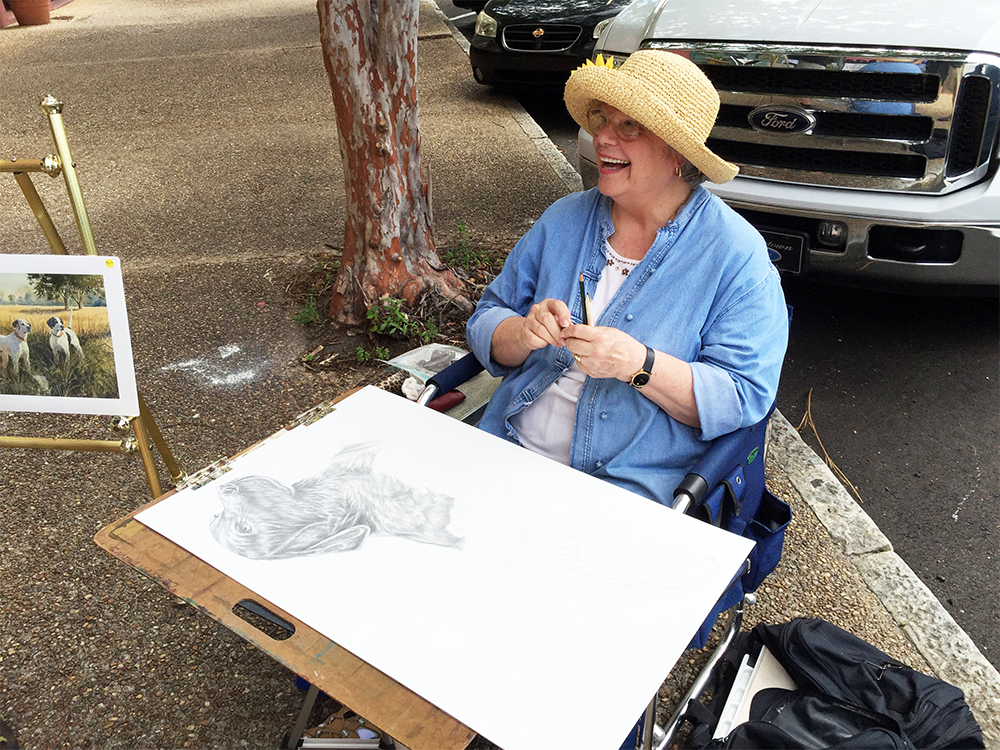 Geri Davis, smiling in a camp chair on the sidewalk with easels and art supplies nearby