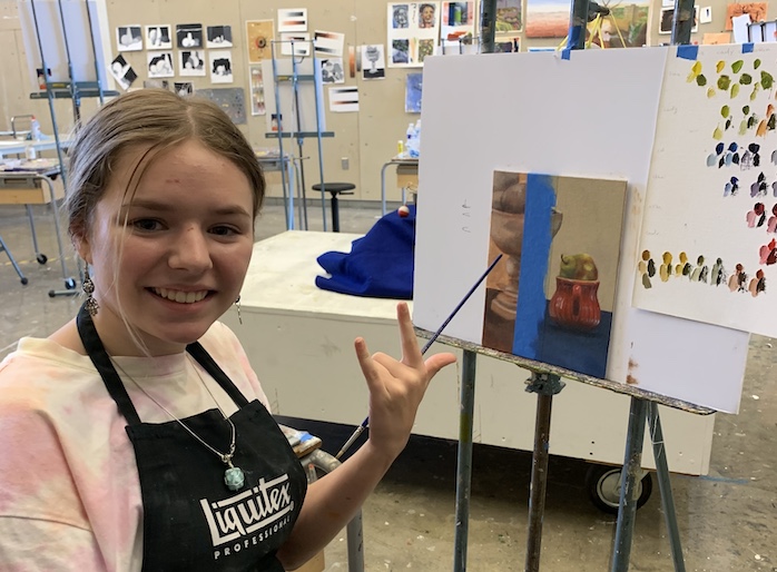 A student in a smock smiling next to her painting