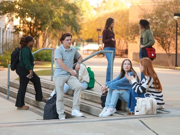 Three students collaborating at a laptop in a bright campus study area, focused and engaged, surrounded by books and modern furniture