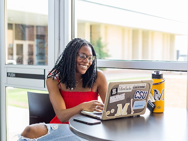 A student happily studying on her personal laptop by a bright window.