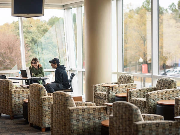 Two students collaborating at a laptop in a bright campus study area, surrounded by modern furniture