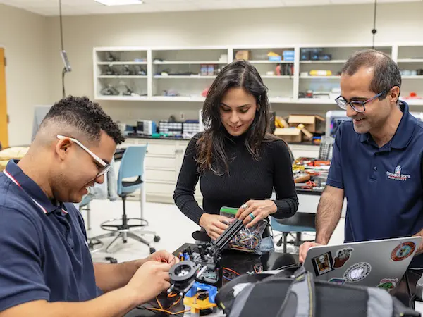 Two students building a robot together in a classroom with a professor overseeing them.