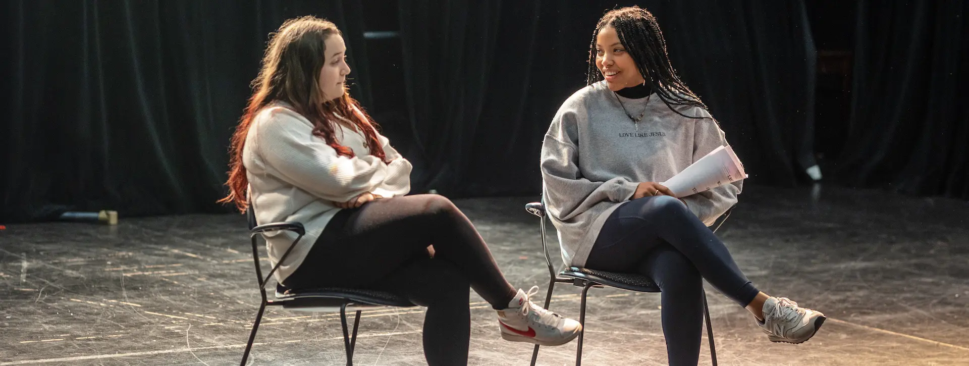 two female students sitting in chairs on stage practicing lines