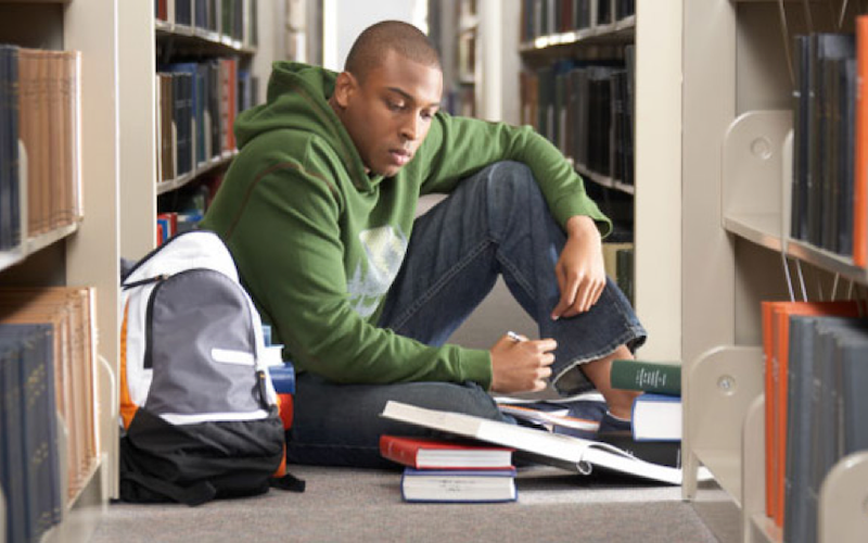 A student in the library, sitting on the floor taking notes next to a stack of books