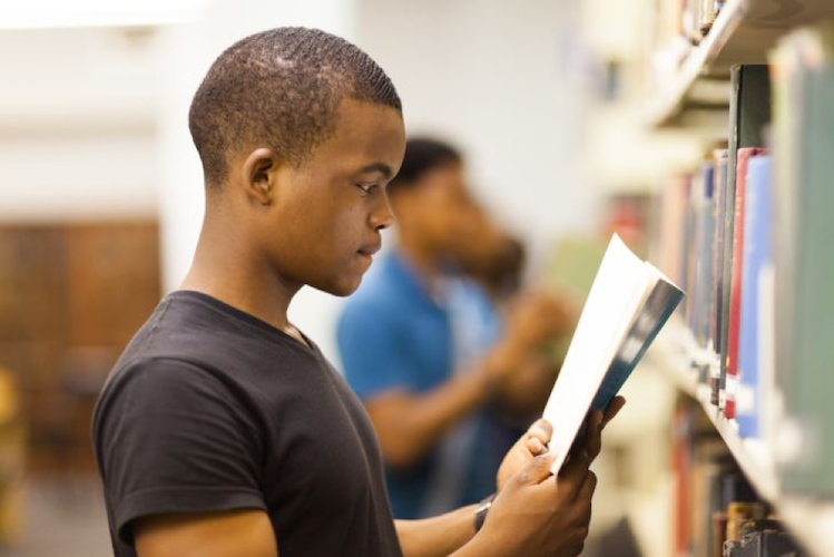A standing student reading next to a library shelf