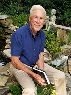 an older man sitting on a rock fountain with plants and a statue behind him
