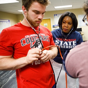 a male student listening to a stethoscope with a female professor standing behind him