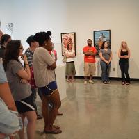 A speaker lecturing in an art gallery with students looking on