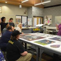 A professor lecturing while students look on, large drawings on a table between them 