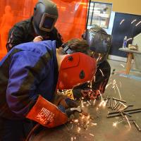 Three individuals working with metal and a torch, all wearing protective gloves and face shields 