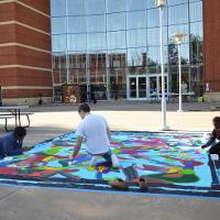 Three people working outside on a large mural lain flat on the ground