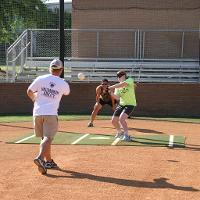 A coach throws a baseball for a blindfolded camper to hit. A camp counselor stands behind the camper, watching both of them.