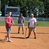 A coach, a camp counselor, and a camper stand together on a baseball field holding baseball bats. 