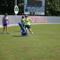 A camp counselor watches a blindfolded camper run past a falling blue training dummy on a baseball field.