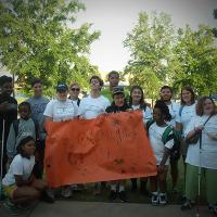 A group of campers stands together to pose for the camera. The campers in the center of the group hold up a large sheet of orange paper with the phrase "Orange Crush" and their names written on it.