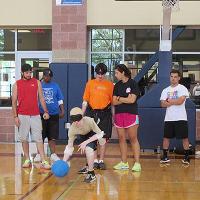A blindfolded camper rolls a ball while several campers, camp counselors, and a coach watch from behind.