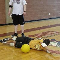 A camper wearing kneepads slides across the gymnasium floor to block a dodgeball while another camper stands behind and watches.