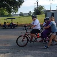 A coach rides a tandem bike with a camper while another coach helps steady the camper on the bike.