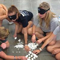 A group of blindfolded campers sort through three different sizes of marshmallows and create shapes with the marshmallows and toothpicks.