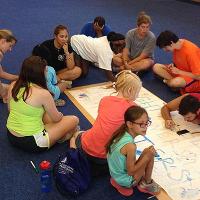 A group of campers sit on the floor around a large white sheet of paper and write on it with markers.