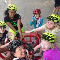 Four campers and a camp counselor wearing helmets ride a multi-person bike, some of them turning to smile for the camera.