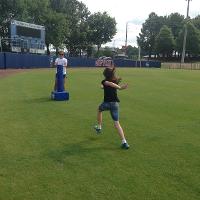 A camp counselor stands behind a blue tackling dummy on a baseball field while a camper runs at the tackling dummy.