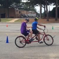 Two campers ride a tandem bike through a parking lot.