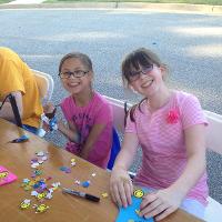 Two campers sit together at a crafting table with foam stickers in front of them and smile for the camera.