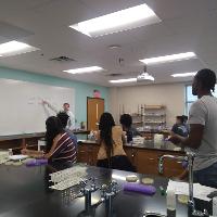 several students paying attention in class to a teacher, a teacher is at the front pointing to a whiteboard