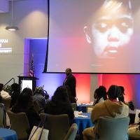 several people watching a speaker on a stage with a photo of a child on a projector screen