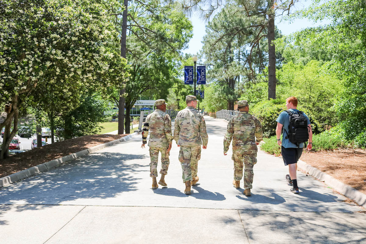Four students walking side by side, three in full military fatigues, one in shorts and a tee shirt