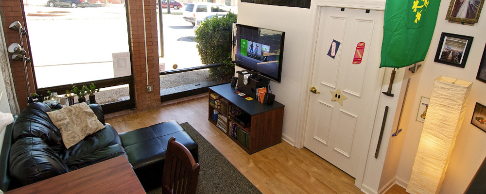 common area with black leather couch, TV on a media console, a table with a single chair, and two large windows