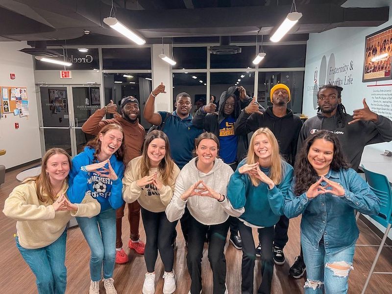 Sorority and fraternity members posing inside, smiling, all making the same hand symbols